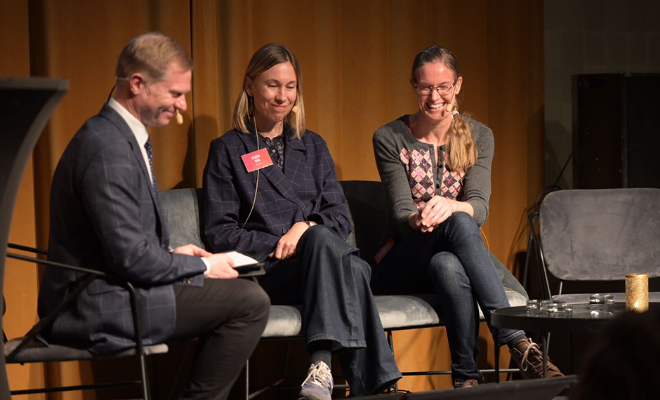 En man och två kvinnor sitter på en scen, tittar på en skärm och skrattar.
